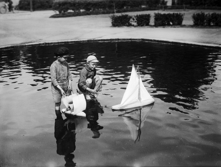 Kaksi nuorta lasta kahluualtaassa, toinen heistä kyykyssä. Kuvassa kaksi valkoista laivaa. Toinen vedessä, toinen lapsen kädessä.