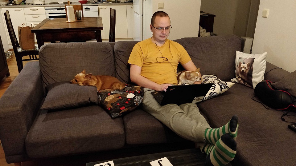 Mies istuu sohvalla ja pitää jalkojaan olohuoneen pöydällä. Hänen sylissään on kannettava tietokone ja korvissa kuulokkeet. Miehen molemmilla puolilla nukkuu pieni koira, joista toinen pitää leukaansa miehen vasemman käden päällä.