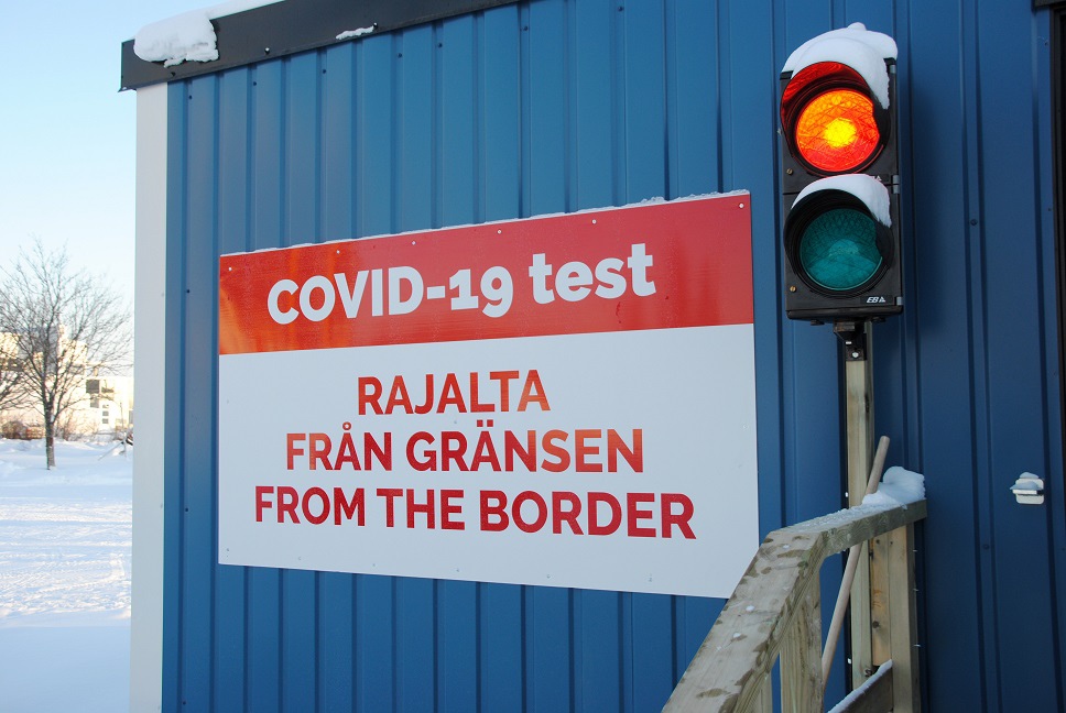 Sininen kontti, jonka seinässä punavalkoinen kyltti tekstillä "COVID-19 test. Rajalta, från gränsen, from the border". Oikealla liikennevalo, jossa palaa punainen valo.
